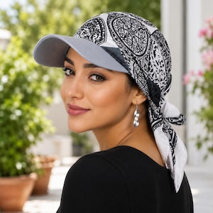 Puede incluir: Un cubrecabezas con una visera gris claro lisa unida a una gorra de tela estampada. La tela presenta un intrincado estampado de estilo damasco en blanco y negro e incluye largas cintas de tela en la parte posterior para anudar.