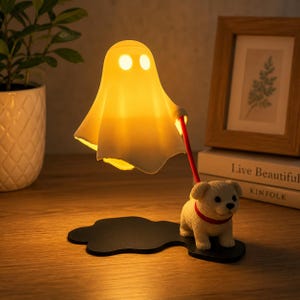 Può includere: Una lampada bianca luminosa a forma di fantasma con due occhi circolari, collegata da un guinzaglio rosso a una piccola statuetta di cucciolo beige. La lampada e il cucciolo sono montati su una base nera a forma di nuvola, emettendo luce.