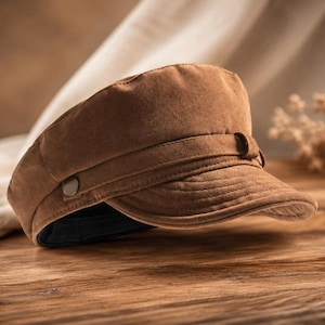 May include: A brown suede-textured military-style cap featuring a flat top, a short curved brim, and a decorative horizontal strap across the front secured by a small metal button on the side. The interior lining is black.