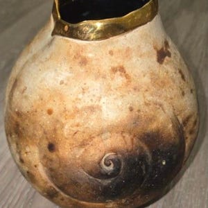 May include: A round ceramic vase with a textured, mottled beige and brown surface featuring a prominent dark brown spiral pattern on the side. The vase has an irregular, wavy rim finished with a metallic gold glaze and a dark interior.