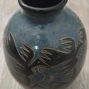 May include: A round ceramic vase with a dark blue and black speckled glaze. The exterior features etched, stylised bird figures in black with fine white outlines. The vase has a smooth, rounded body and a short, dark-rimmed circular opening.