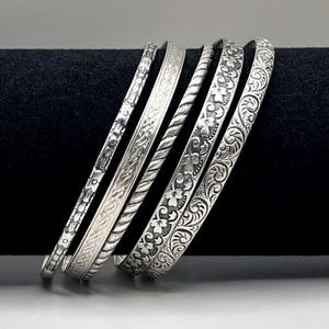 May include: A set of five silver-toned metal bangles featuring various embossed patterns, including floral motifs, intricate scrollwork, and a twisted rope design. Each bracelet has a distinct texture and width, showcasing detailed metallic craftsmanship.