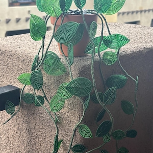 May include: A decorative trailing plant sculpture crafted from small green beads, featuring numerous leaf-shaped segments attached to flexible stems. The beaded plant is potted in a simple terracotta-coloured container filled with dark pebbles.