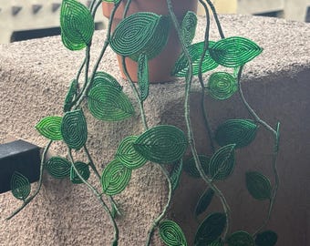 Puede incluir: Una escultura de planta colgante hecha de pequeñas cuentas verdes, con numerosos segmentos en forma de hoja unidos a tallos flexibles. La planta de cuentas está colocada en una maceta de color terracota llena de guijarros oscuros.