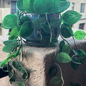 May include: A decorative plant sculpture featuring numerous vibrant green beaded leaves arranged on thin, flexible stems. The beaded foliage is contained within a smooth, dark blue ceramic pot that sits on a matching saucer, creating a lifelike appearance.