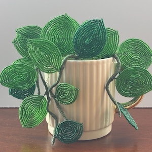 May include: A cream-colored, vertically ribbed ceramic mug containing a decorative plant sculpture made of green glass beads. The plant features multiple wire-stemmed branches with circular and oval-shaped leaves crafted from small, shiny green beads.