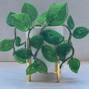 May include: A decorative plant sculpture featuring vibrant green beaded leaves on flexible wire stems. The plant is housed in a smooth white cylindrical ceramic pot, which is supported by a polished gold-toned metal stand with four vertical legs.