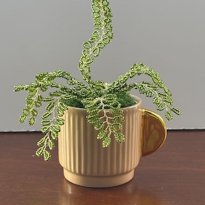 May include: A decorative plant sculpture made of green glass beads on thin wire stems, arranged in a light beige ceramic mug with vertical ridges and a metallic gold handle. The plant is set into a green moss-like base inside the mug.