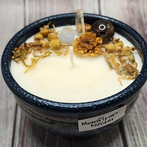 May include: A blue ceramic bowl candle with a white wax base. The candle is decorated with dried flowers, crystals, and a label that reads "Moonflower Nectar".