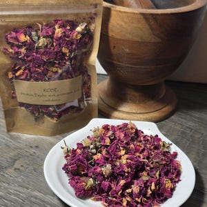 May include: Dried rose petals in a clear bag with a brown label that reads "ROSE". A white plate holds a pile of rose petals. A wooden mortar and pestle are in the background. The label also includes the words "Intuition, Psychic work, protection, and confidence."