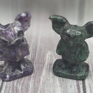 May include: Two carved stone figurines. One is amethyst, with purple and white hues. The other is green with dark speckles. Both have large ears and a seated pose, resembling a stylized creature.