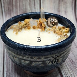May include: A blue ceramic bowl candle with a white wax top. The candle has a single wick and is decorated with dried flowers, crystals, and a small stone. The bowl has a blue and white glaze with a pattern on the side.