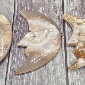 May include: Three crescent moon-shaped carvings made of agate stone. The moons have a natural, marbled appearance with shades of beige, cream, and brown. The carvings are displayed on a wooden surface.