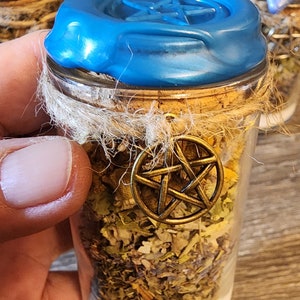 May include: A small glass jar filled with dried herbs and a white powder base. The jar is sealed with a cork and blue wax, and adorned with a gold-colored pentacle charm. The jar is held in a hand.