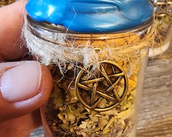 May include: A small glass jar filled with dried herbs and a white powder base. The jar is sealed with a cork and blue wax, and adorned with a gold-colored pentacle charm. The jar is held in a hand.