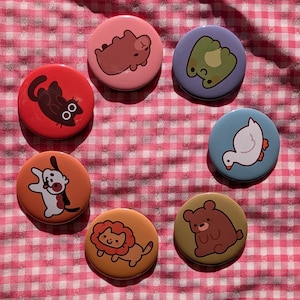 May include: Seven circular pin-back badges featuring cartoon illustrations of animals. The designs include a brown bear on pink, a green frog on purple, a white duck on blue, a brown bear on olive, a lion on orange, a dog on peach, and a black cat on red.