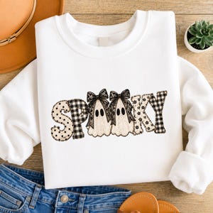 Könnte beinhalten: Ein weißes Langarm-Sweatshirt mit dem Wort SPOOKY in einer dekorativen Schriftart. Die Buchstaben sind mit schwarz-weißen Mustern wie Punkten, Vichy-Karos und Blumenmustern gefüllt. Zwei Geisterfiguren mit schwarzen Schleifen sind zentriert.