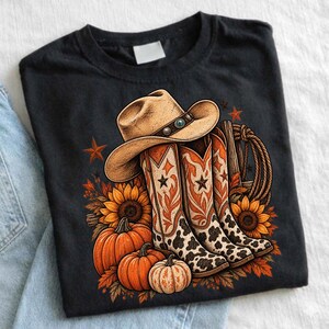 Könnte beinhalten: Ein schwarzes T-Shirt mit einem Grafikdruck eines beigen Cowboyhuts, der auf einem Paar Westernstiefeln mit Kuhfleckenmuster und orangefarbenen Verzierungen ruht, umgeben von orangefarbenen Kürbissen, Sonnenblumen, Herbstblättern, einem Seil und Sternen.