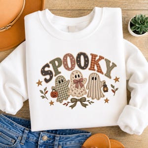 Könnte beinhalten: Ein weißes Sweatshirt mit Rundhalsausschnitt und dem Wort SPOOKY in gemusterten, mehrfarbigen Stoffbuchstaben. Unter dem Text befinden sich drei Geister: einer mit grünem Karomuster und Kürbis, einer mit Blumenmuster und Schleife sowie einer mit Streifen und Laterne. Eine Zierschleife und kleine Sterne umgeben die Geister.