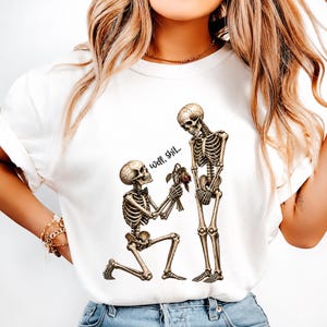 May include: A white t-shirt featuring a graphic of two skeletons. One skeleton is kneeling and wearing a small black bow tie, offering a wilted rose to the other standing skeleton. The text "Well, Shit..." is printed in black script above the rose.
