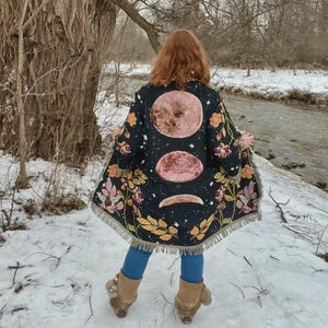 Op de afbeelding: Een zwart geweven vest met een maanfase-afbeelding met drie maancycli, omringd door kleurrijke bloemen- en bladpatronen. Het kledingstuk heeft een gefranjerde zoom en lange mouwen, met een gedetailleerd ontwerp in wandtapijstijl op de achterkant.