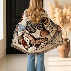 May include: A cream-coloured woven cardigan featuring a large, multi-coloured butterfly print across the back and sleeves. The garment has long sleeves, an open front, and a frayed fringe hemline. It is paired with light blue denim jeans.