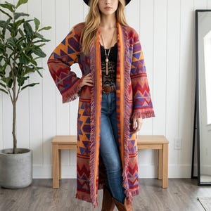 May include: A long, open-front cardigan featuring a vibrant geometric pattern in shades of orange, red, purple, and blue. The garment includes long sleeves, a straight hemline, and decorative fringe trim along the front edges and sleeve cuffs.