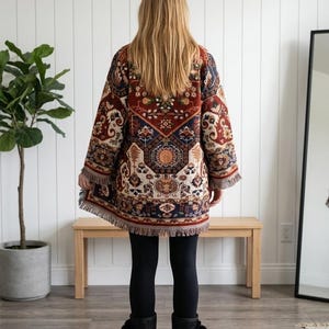 May include: A long-sleeved jacket featuring a multi-coloured tapestry pattern in shades of deep red, navy blue, and cream. The garment has a straight hemline finished with a fringe trim, long sleeves, and a loose fit, worn over black leggings.
