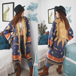 Peut inclure: Un long gilet ouvert orné d'un motif céleste avec un grand visage de soleil dans le dos, des croissants de lune, des étoiles et des planètes dans des tons bleu, or et orange. Le vêtement a des manches longues, un ourlet à franges et une bordure claire.