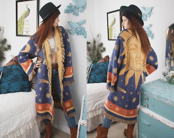May include: A long, open-front cardigan featuring a celestial print with a large sun face on the back, crescent moons, stars, and planets in shades of blue, gold, and orange. The garment has long sleeves, a fringed hem, and a contrasting light-colored trim.