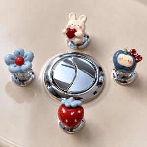 May include: A set of four decorative ceramic knobs featuring a white rabbit holding a red heart, a light blue flower with a red center, a red strawberry, and a blue apple with a white face and red bow, all mounted on circular chrome bases.