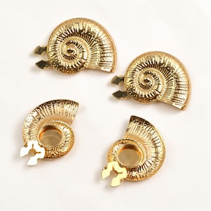 Pode incluir: Quatro botões de metal dourado em forma de conchas de nautilo. Dois botões apresentam uma superfície superior texturizada em espiral com linhas radiais, enquanto os outros dois exibem um centro liso, circular e rebaixado.
