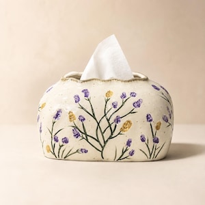 May include: An oval ceramic tissue box cover with a cream-coloured base and a speckled finish. The exterior is decorated with hand-painted green stems featuring small purple and yellow flower buds. A single white tissue protrudes from the top opening.