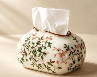 May include: A white ceramic tissue box cover with a rounded, oblong shape, featuring a delicate botanical pattern of green leaves and pink flowers. A single white paper tissue is pulled through the top opening of the container.