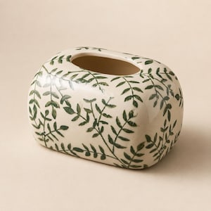May include: A rectangular ceramic tissue box cover with rounded corners, featuring a cream-colored base decorated with a repeating pattern of dark green hand-painted leafy vine sprigs across the exterior surface and a large oval opening.