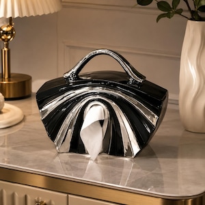 May include: A black and silver ceramic tissue box holder shaped like a handbag with a curved handle. The design features a fan-like pattern with alternating black and metallic silver sections, and a central opening dispensing a single white tissue.