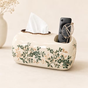 May include: A cream-colored ceramic tissue box holder with a brown-rimmed opening for tissues and a separate side compartment. The exterior features a repeating pattern of green and light brown floral vines. A black remote and glasses are inside.