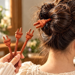 May include: A set of reddish-brown wooden hair sticks, each featuring a decorative top shaped like a two-prong electrical plug. The sticks have a smooth, polished finish and a long, slender stem designed to secure hair in a bun or updo style.