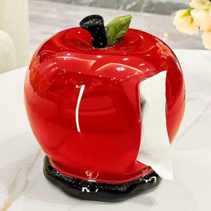May include: A glossy red apple-shaped tissue dispenser with a black base and a black stem featuring a single green leaf. A white tissue is pulled through a vertical opening on the side of the round, shiny ceramic-like container.
