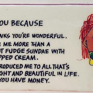 May include: A rectangular, white novelty item with the text "I LOVE YOU BECAUSE" and humorous reasons. A cartoon figure with red hair and a yellow dress with green polka dots is on the right.