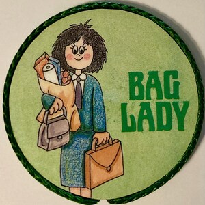 May include: A circular pin featuring a cartoon illustration of a person with dark hair wearing a blue suit, holding a brown paper bag filled with items, a small gray handbag, and a brown briefcase. The text BAG LADY is printed in green.