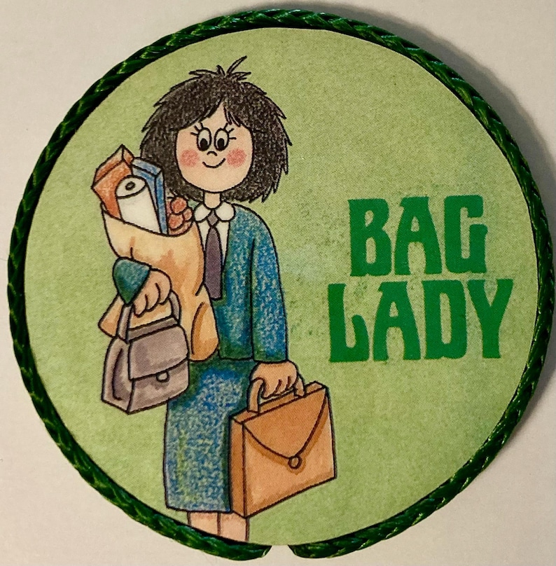 May include: A circular pin featuring a cartoon illustration of a person with dark hair wearing a blue suit, holding a brown paper bag filled with items, a small gray handbag, and a brown briefcase. The text BAG LADY is printed in green.