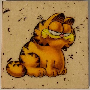 Pu&ograve; includere: Un sottobicchiere quadrato con un'immagine a cartoni animati di Garfield il gatto. Garfield &egrave; arancione con strisce nere, un naso rosa e un'espressione scontrosa. Lo sfondo &egrave; beige chiaro con macchie scure.