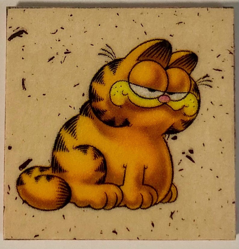 Pu&ograve; includere: Un sottobicchiere quadrato con un'immagine a cartoni animati di Garfield il gatto. Garfield &egrave; arancione con strisce nere, un naso rosa e un'espressione scontrosa. Lo sfondo &egrave; beige chiaro con macchie scure.