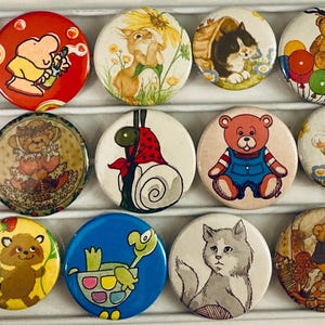 May include: A collection of colorful vintage pinback buttons featuring various cartoon animals and characters. Designs include a turtle, cat, bear, koala, and raccoon. The buttons are round and have a metal backing.