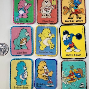 Peut inclure: Neuf cartes colorées représentant des Care Bears et des Schtroumpfs. Les cartes sont rectangulaires et ont une bordure blanche avec un bord cousu bleu, rose, jaune ou marron. Chaque carte présente un personnage différent et un nom. Les personnages incluent Cheer Bear, Friend Bear, Cowboy Smurf, Wish Bear, Funshine Bear, Hefty Smurf, Bedtime Bear, Birthday Bear et Skier Smurf.