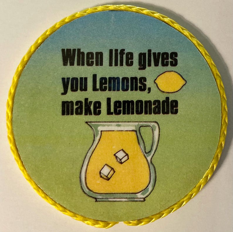May include: A circular coaster with a yellow braided border. The center features a blue and green gradient background with the black text "When life gives you Lemons, make Lemonade," a small yellow lemon icon, and a pitcher of yellow lemonade.
