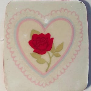 May include: A square, off-white decorative tile featuring a red rose with green leaves inside a pink and blue heart. The heart is surrounded by a scalloped border. The tile has rounded corners.