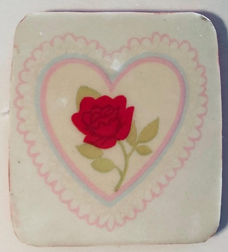 May include: A square, off-white decorative tile featuring a red rose with green leaves inside a pink and blue heart. The heart is surrounded by a scalloped border. The tile has rounded corners.