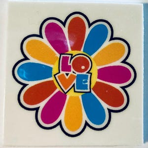 May include: A colorful flower sticker with a black outline. The flower petals are in shades of orange, pink, blue, and yellow. The word "LOVE" is in the center of the flower in a retro font. The sticker is on a white square.